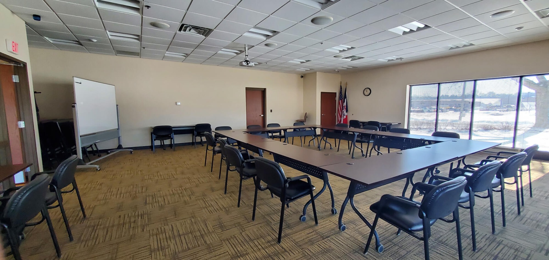 Large, bright meeting room with a white board in the corner and a couple of small desk arrangements against the walls. Four long tables face one another with 16 chairs surrounding it.